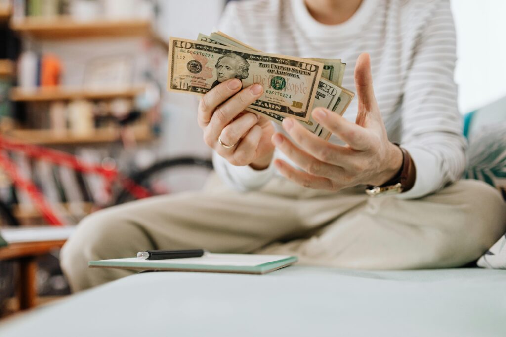 Man wearing casual clothes counts dollar bills while sitting on a sofa indoors.