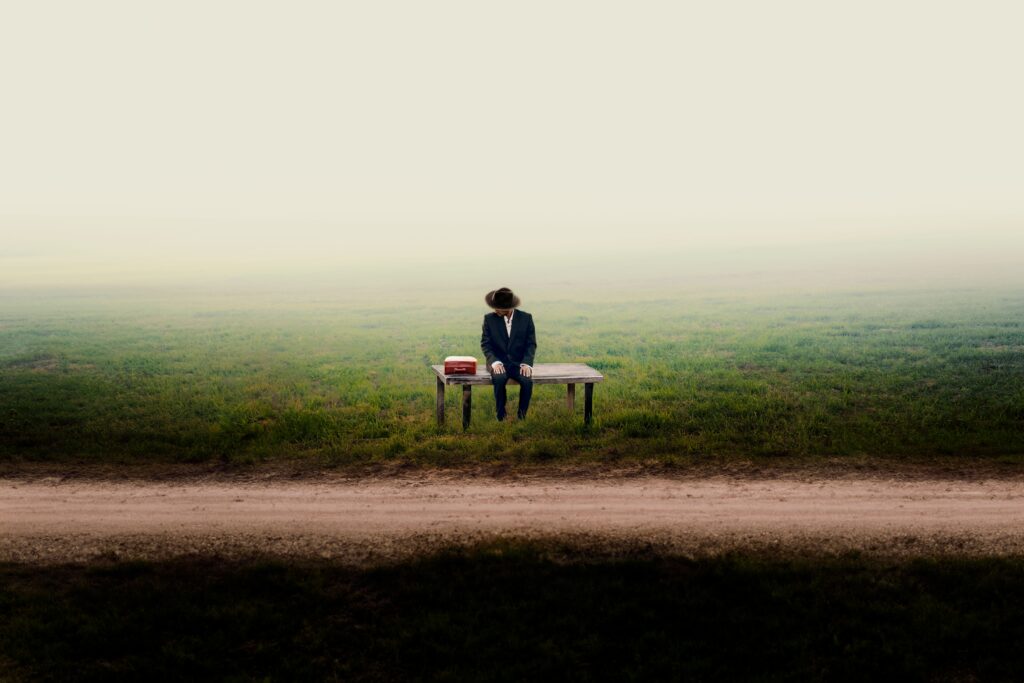 A man in a suit sits alone on a wooden table in a foggy field, depicting solitude and introspection.